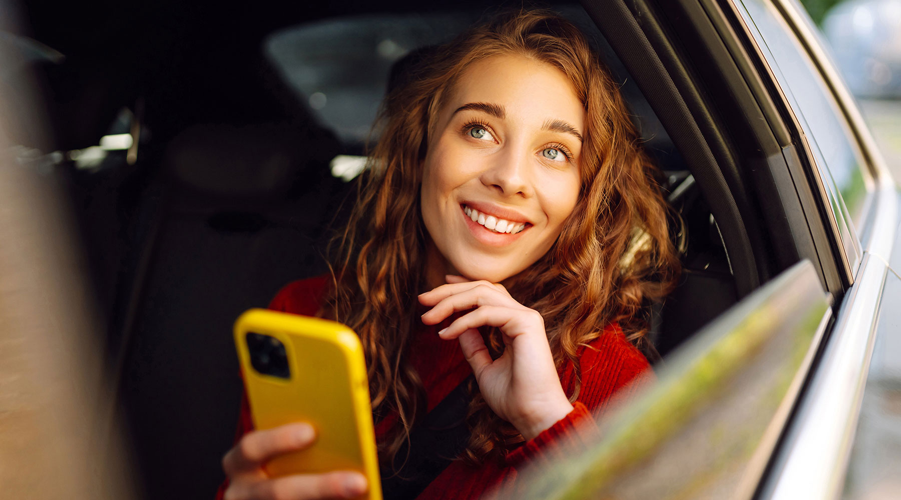 Junge Frau sitzt im Auto und schaut aus dem Fenster, dabei hält sie ein Smartphone mit gelber Hülle in der Hand.