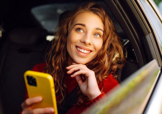 Junge Frau sitzt im Auto und schaut aus dem Fenster, dabei hält sie ein Smartphone mit gelber Hülle in der Hand.
