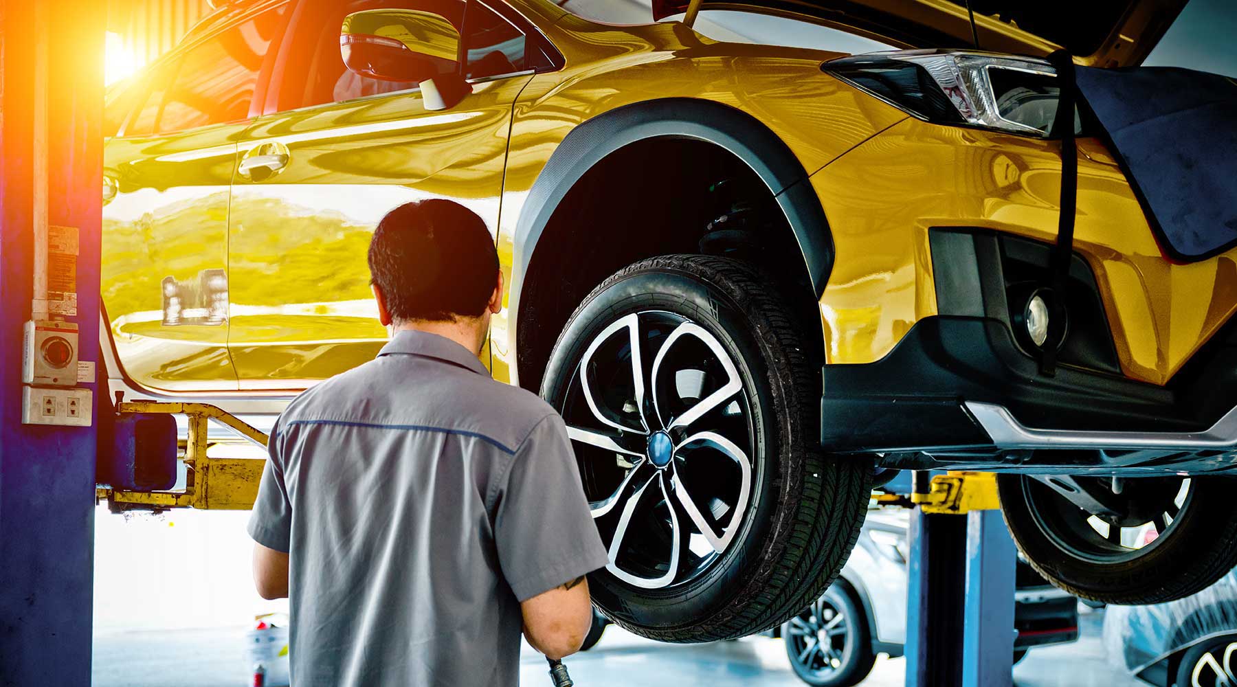 Mann im grauen Shirt schraubt an einem gelben Auto auf einer Hebebühne.