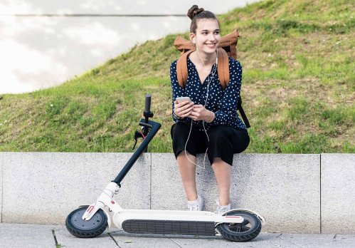 Junge Frau sitzt auf einer niedrigen Mauer. Vor ihr liegt ein E-Scooter.