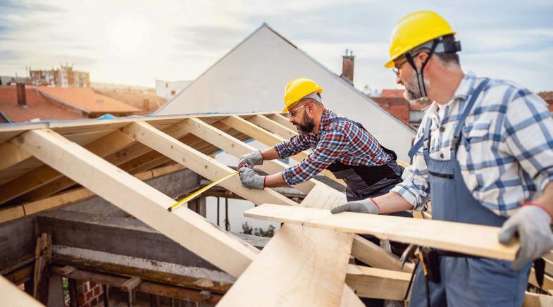 Zwei Handwerker mit Helmen stehen auf einem Haus und reparieren den Dachstuhl mit Holzbalken