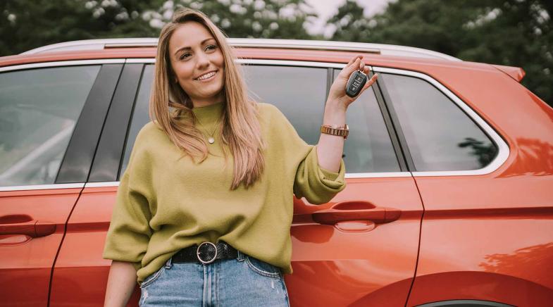 Junge Frau steht vor ihrem Auto und hält den Autoschlüssel hoch.