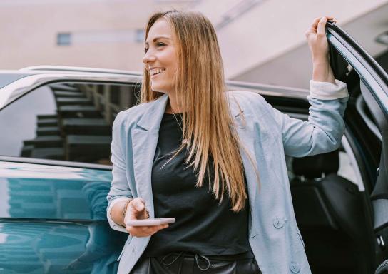 Junge Frau steigt aus dem Auto und hält ihr Smartphone locker in der Hand.