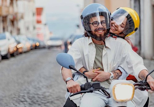 Pärchen fährt fröhlich auf einem Moped über eine Pflasterstraße.