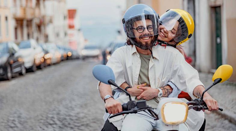 Pärchen fährt fröhlich auf einem Moped über eine Pflasterstraße.