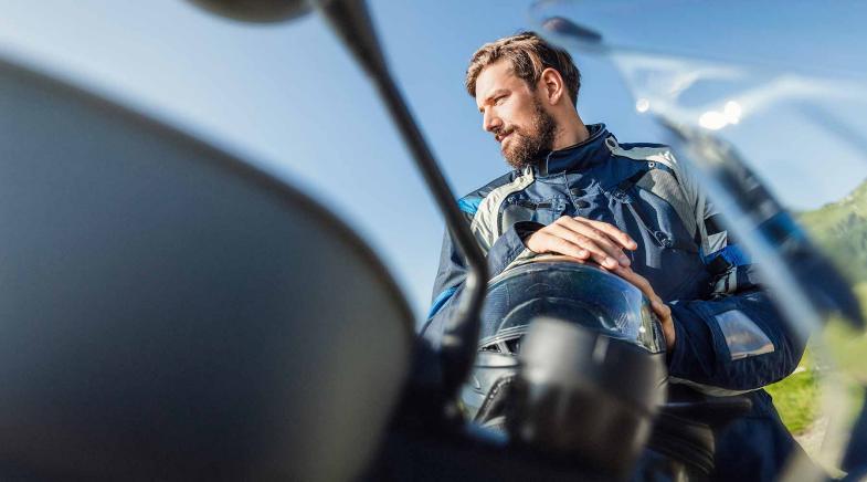 Mann in Motorradschutzkleidung mit Helm in der Hand spiegelt sich im Lack seines Motorrads.