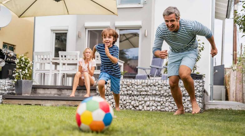 Ein Vater spielt mit seinem Sohn im Garten Fußball während die Mutter auf der Terrasse sitzt und zuschaut.