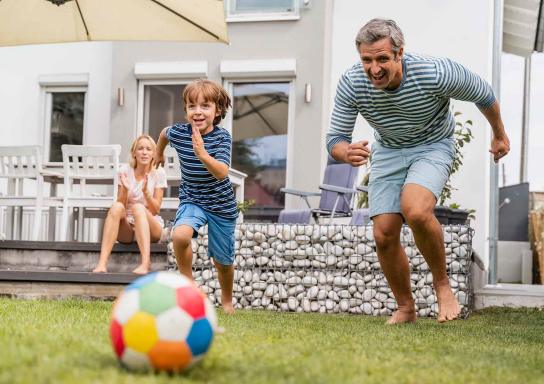 Ein Vater spielt mit seinem Sohn im Garten Fußball während die Mutter auf der Terrasse sitzt und zuschaut.