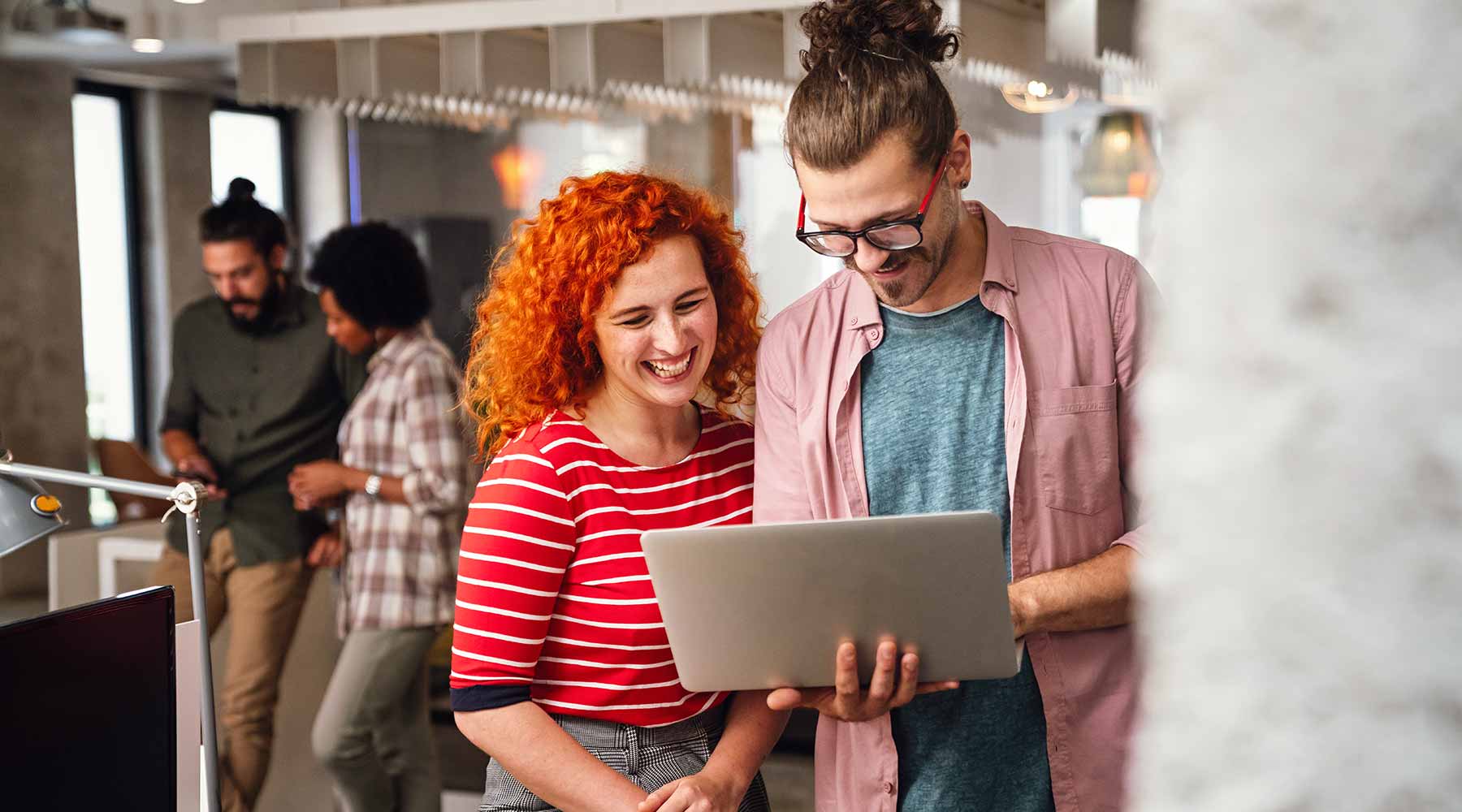 Studentin und Student schauen lachend auf eine Laptop, den der Student auf der Hand hält.