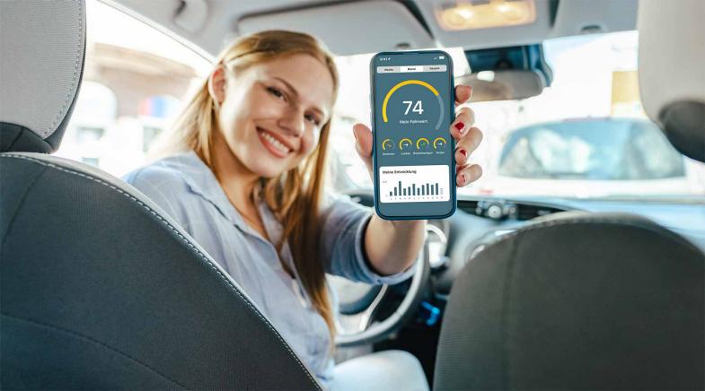 Junge Frau sitzt im Auto, schaut nach hinten und hält ein Handy hoch auf dem ein Screen der HUK-Telematik App zu sehen ist.