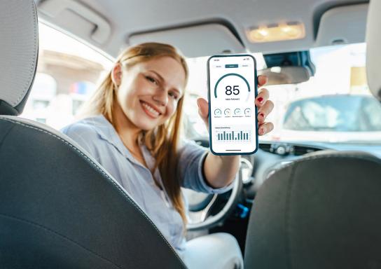 Eine junge Frau sitzt in ihrem Auto und zeigt ihr Smartphone in die Kamera. Auf dem Smartphone ist die App Meine Auto der HUK-COBURG zu sehen.