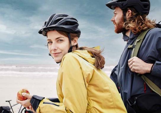 Junges Paar in Regenjacken und mit Helmen schaut am Strand neben ihren Fahrrädern aufs Meer.