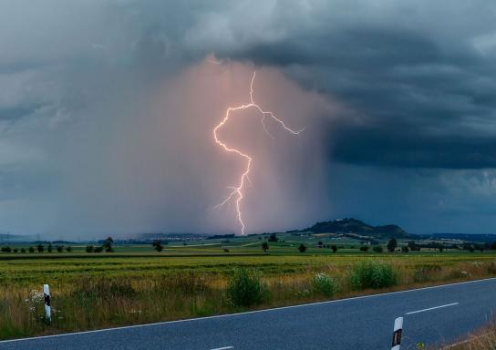 Blitz zuckt aus einer dunklen Unwetterwolke am Horizont neben einem Hügel in den Boden.