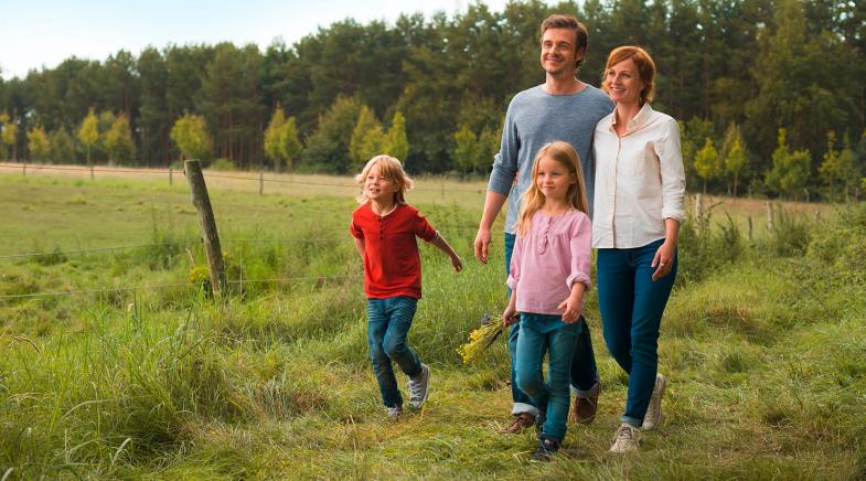 Familie mit zwei Kindern geht am Rand eines Feldes vor einem Wald im Gras spazieren