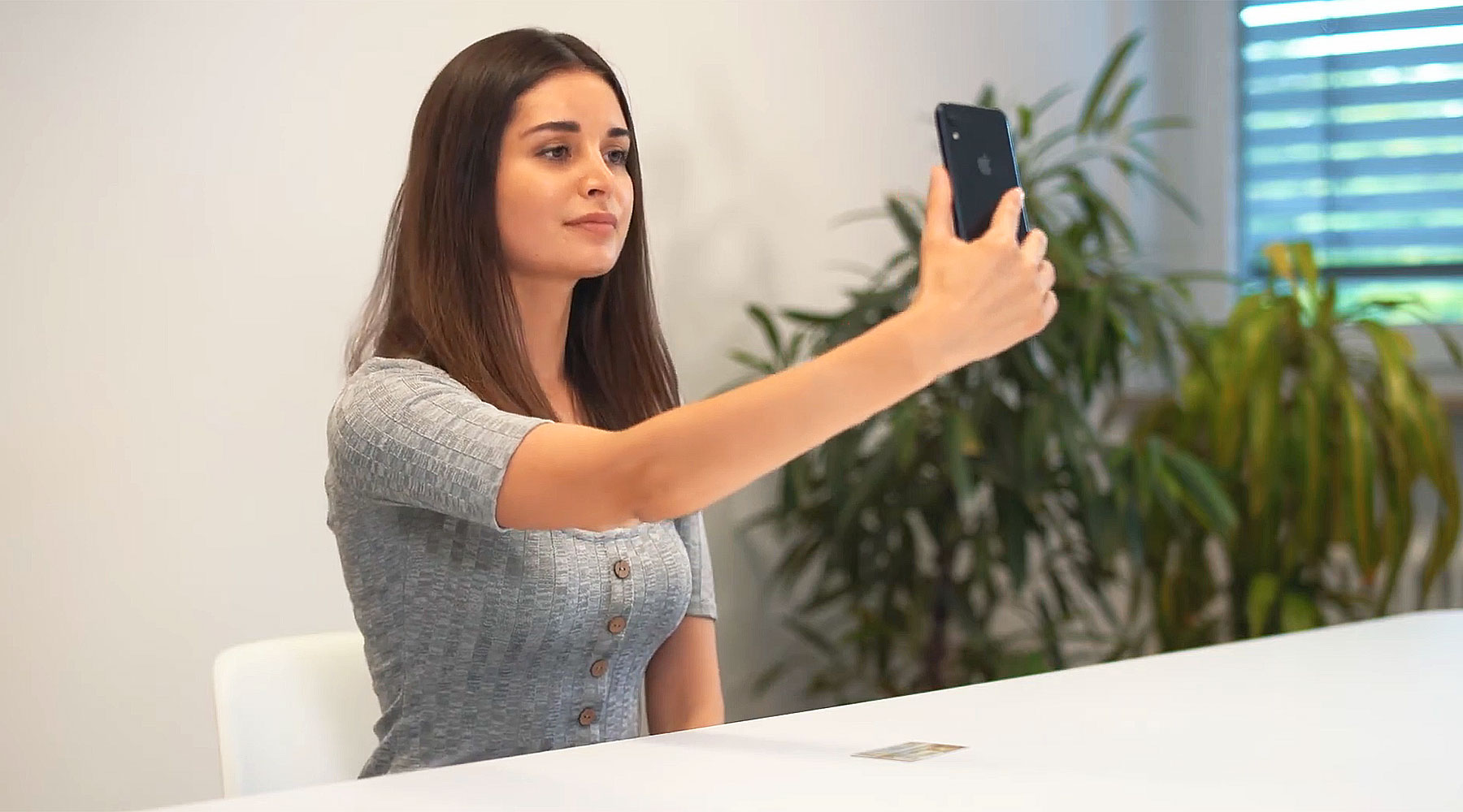 Eine junge Frau sitzt an einem Tisch und hält ein Smartphone vor sich, als ob sie ein Selfie oder ein Foto machen würde. Im Hintergrund befinden sich Pflanzen und ein Fenster mit Jalousien. Auf dem Tisch liegt ein Ausweis.